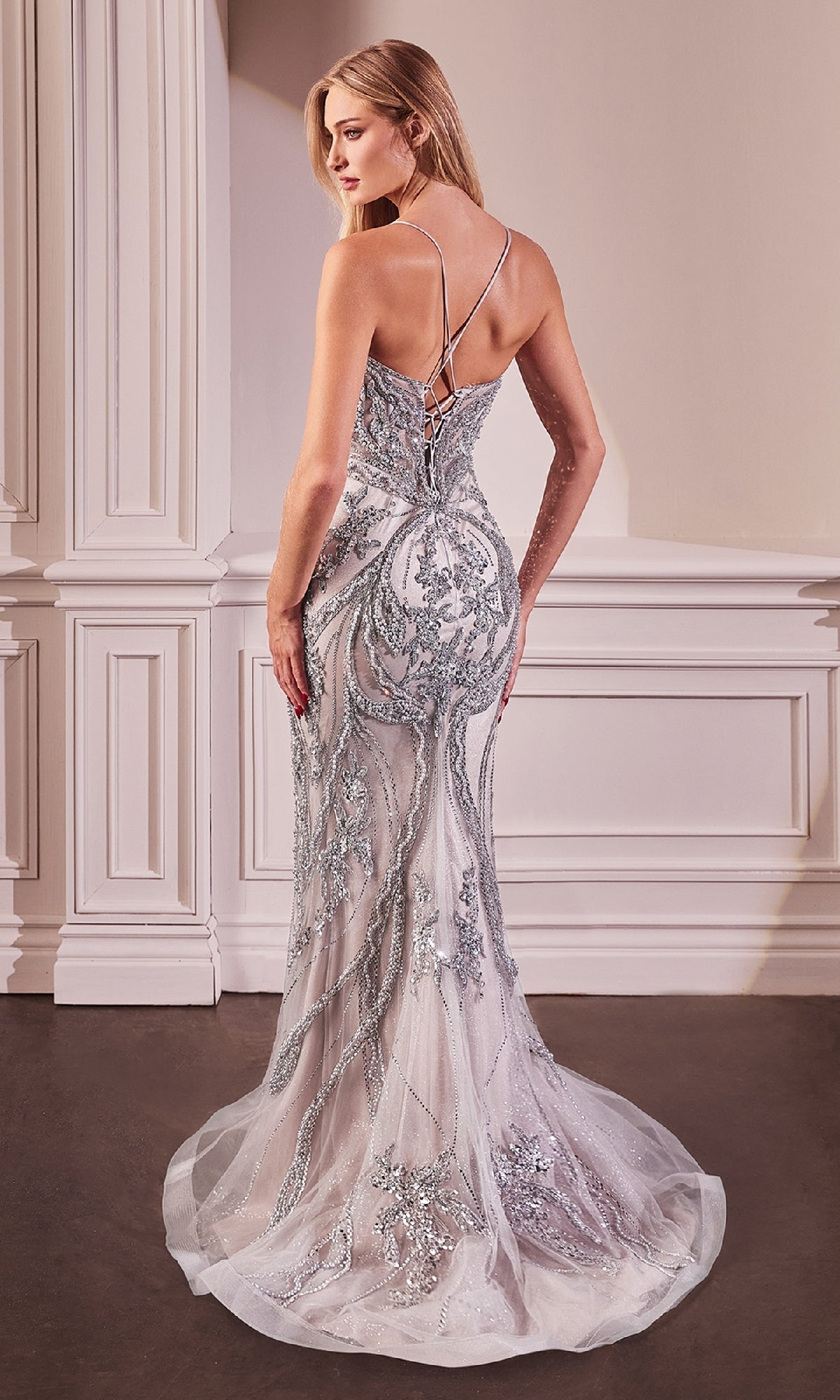 A woman stands in a room wearing the Ladivine CC0240, a long beaded silver prom dress with a lace-up back and trailing hem.